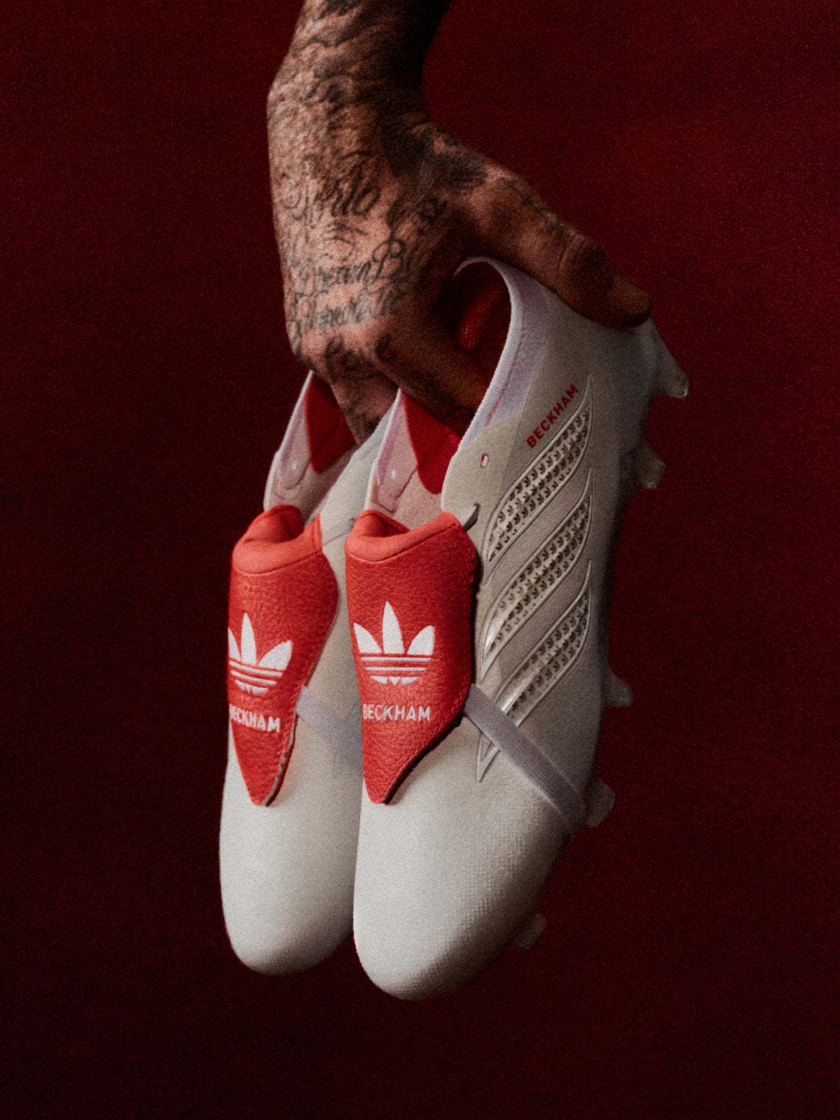 White adidas Predator David Beckham soccer cleats with red fold-over tongues, held by a tattooed hand against a dark red background.
