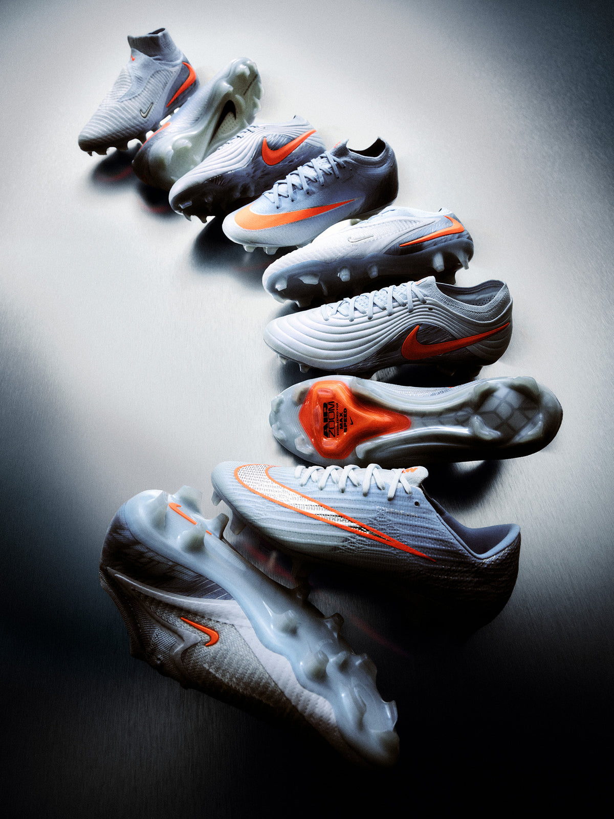 Nike United soccer cleats in silver and light gray with orange Swooshes, arranged diagonally against a dark background.
